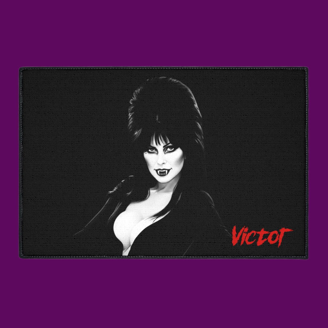 Custom digital illustration of Elvira, Mistress of the Dark, on a plush rectangular bath mat. She is shown in her iconic gothic black dress and beehive hair with a vampire-themed background