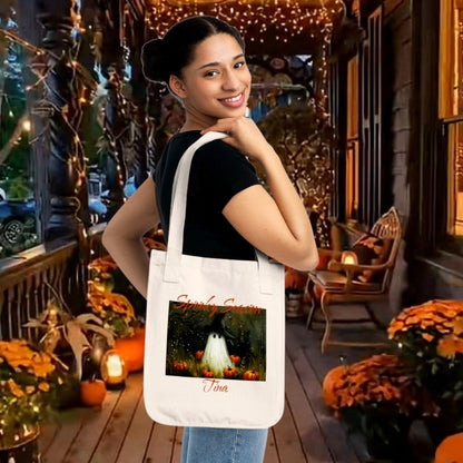 A off white 100% organic cotton tote bag featuring a Halloween-themed illustration of a ghost in a witch hat, surrounded by pumpkins modeled on a lovely fall shopper. Personalize yours in The Fright Factory New Orleans horror gift shop.