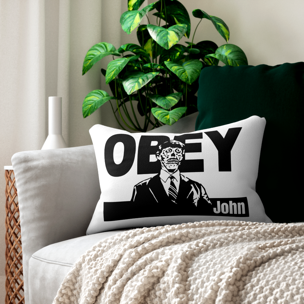 A modern white decorative pillow with black 'OBEY' text inspired by 'They Live' styled on a dark grey sofa. Edgy, minimalist horror movie home decor.